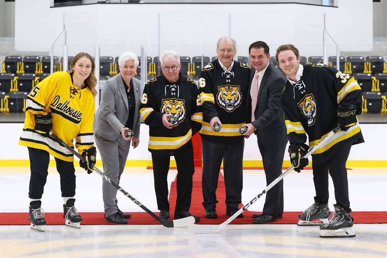 Six people stand on the ice inside a hockey arena: two students in jerseys holding hockey sticks flank four individuals holding pucks, two wearing jerseys and two in suits.