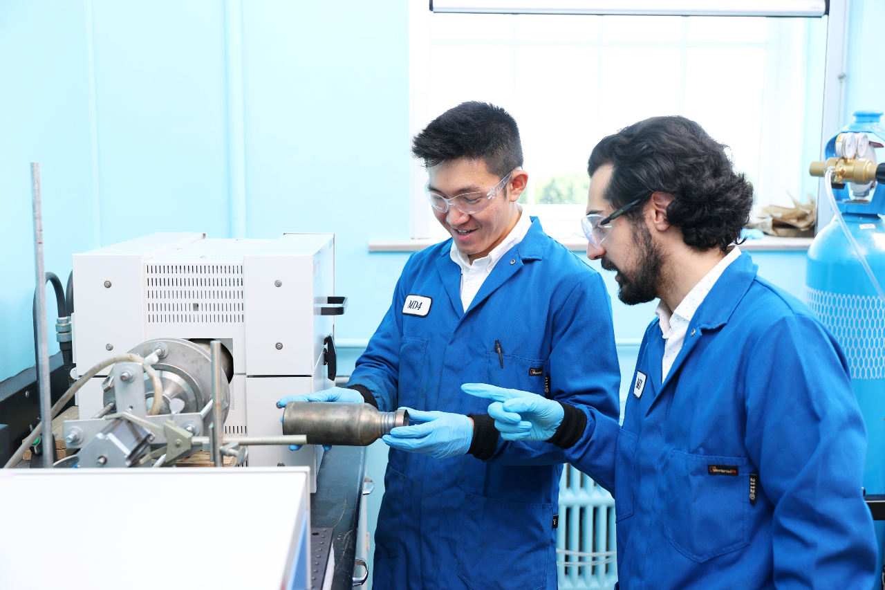 Two men in blue lab coats and gloves examine a metal object