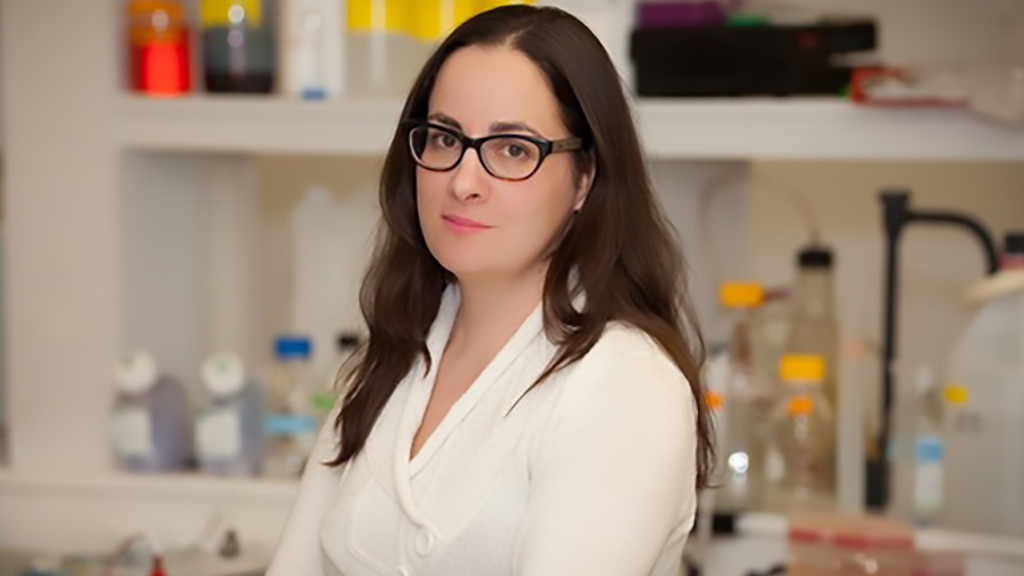 A woman with long brown hair wearing glasses and a white shirt stands in a lab