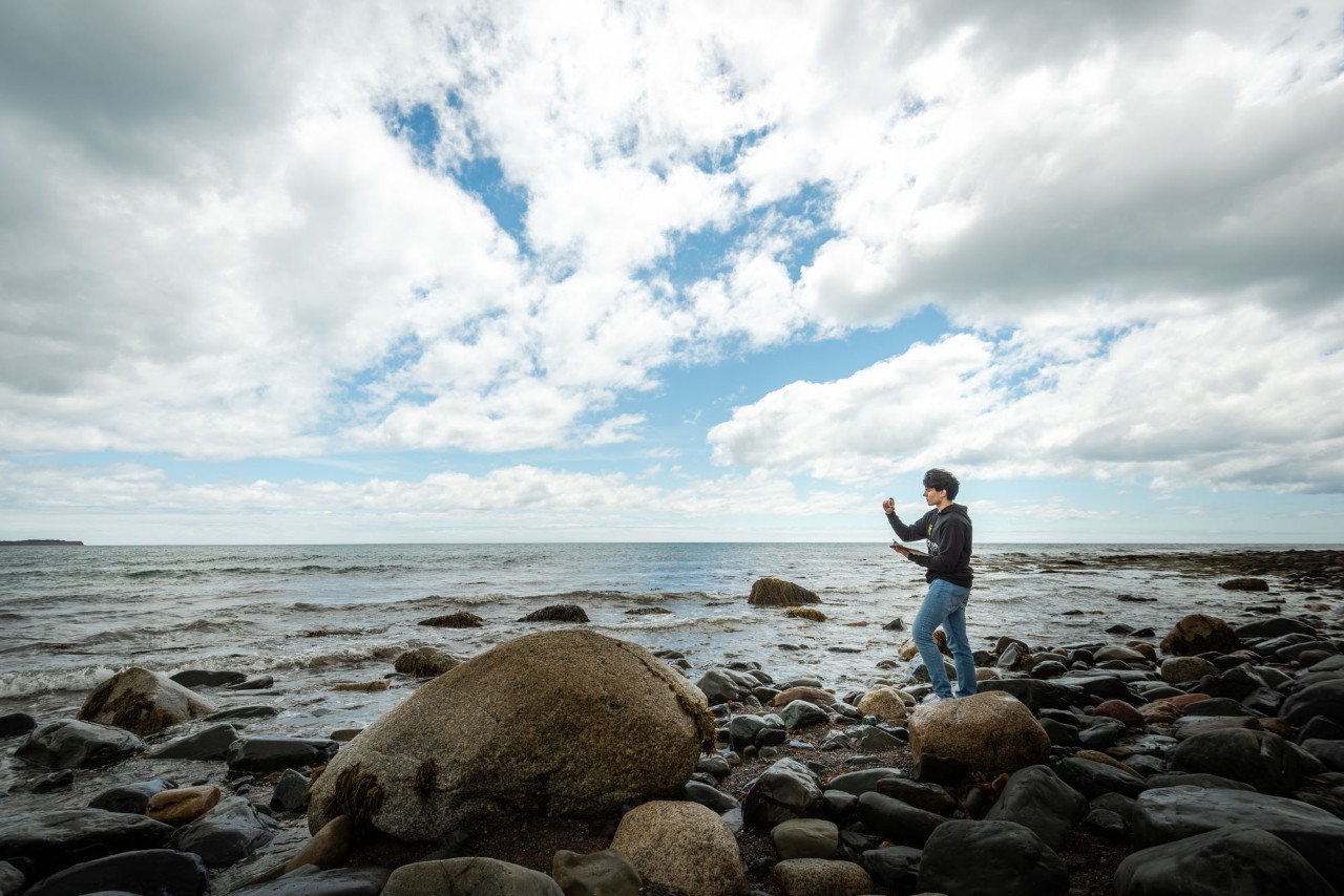 A man wearing a black sweater stands on a rocky beach looking to the ocean