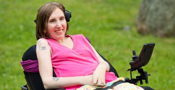 A thin white woman wearing a pink sleeveless shirt sits in a motorized wheelchair, smiling at the camera with her hands clasped in her lap.