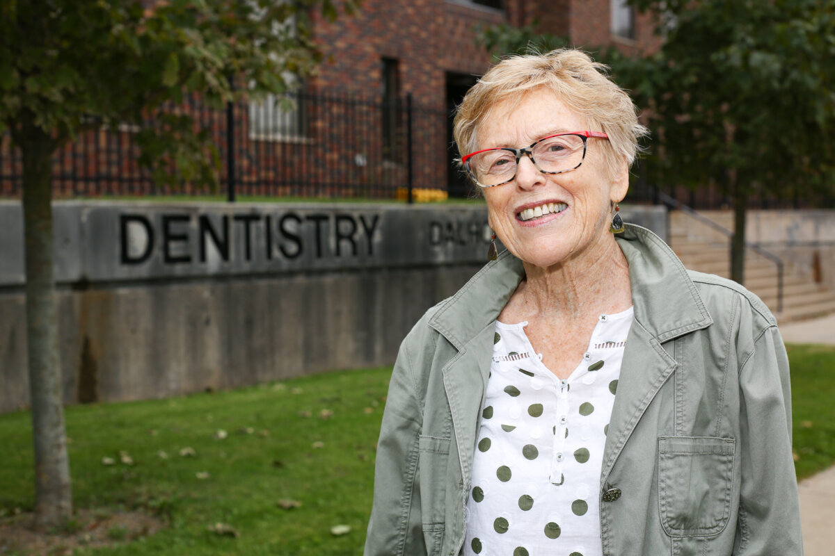 A woman wearing glasses smiles in front of a brick building with a stone sign that reads 'Dentistry.'