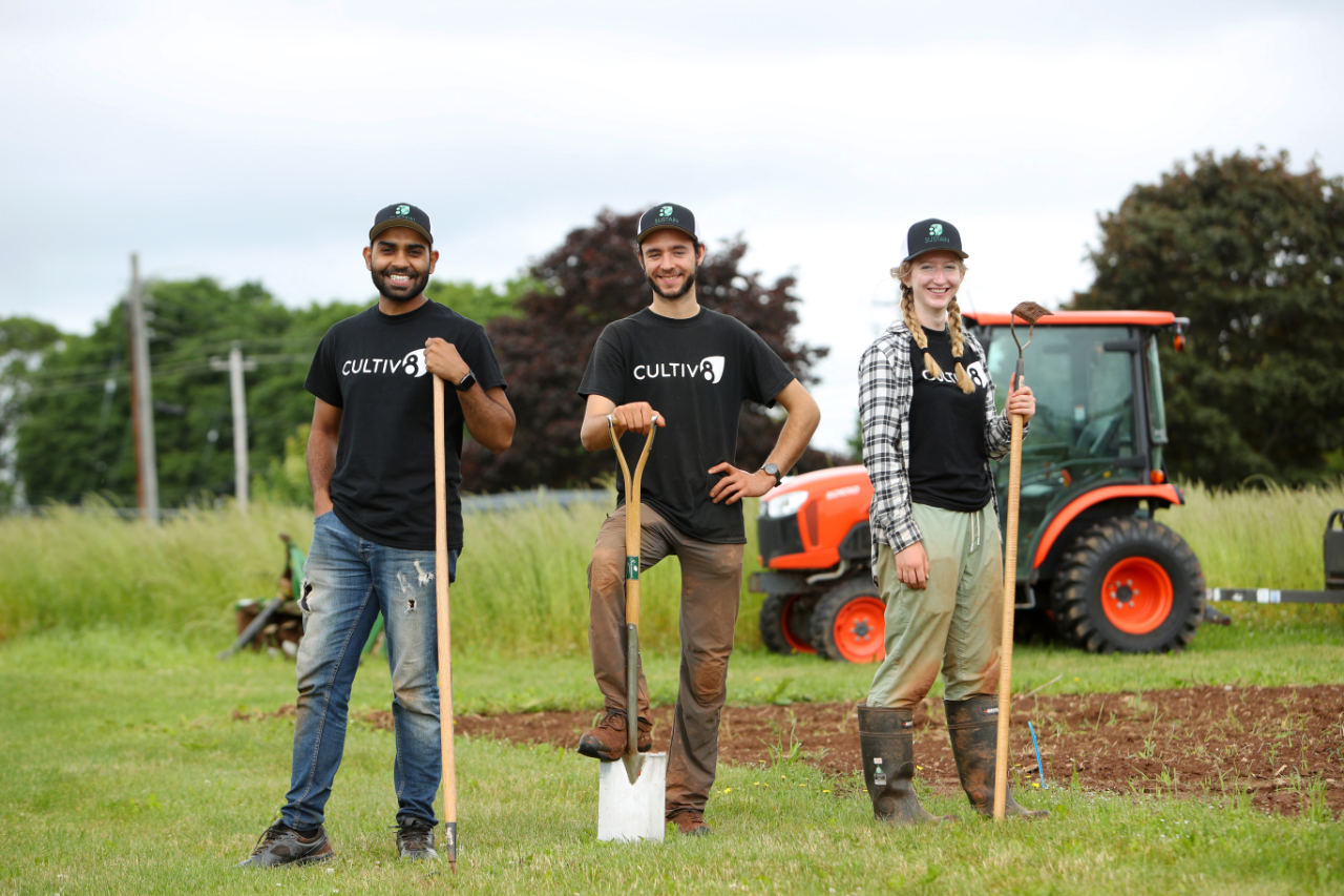 Three people stand on shovels in a field with a tractor in the background