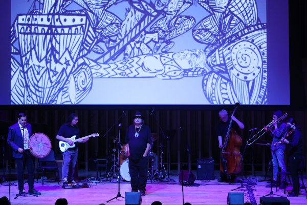 A group of musicians standing on a stage with a screen displaying indigenous art