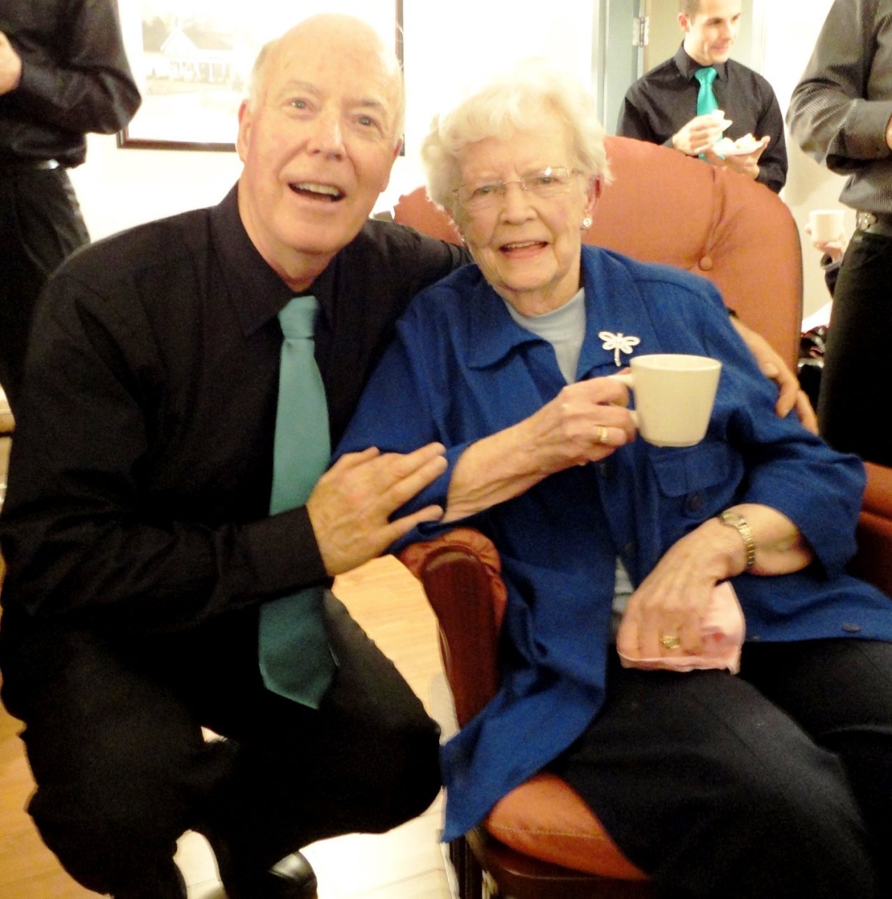 A man wearing a black shirt and green tie squats next to a seatd woman wearing a blue jacket holding a much