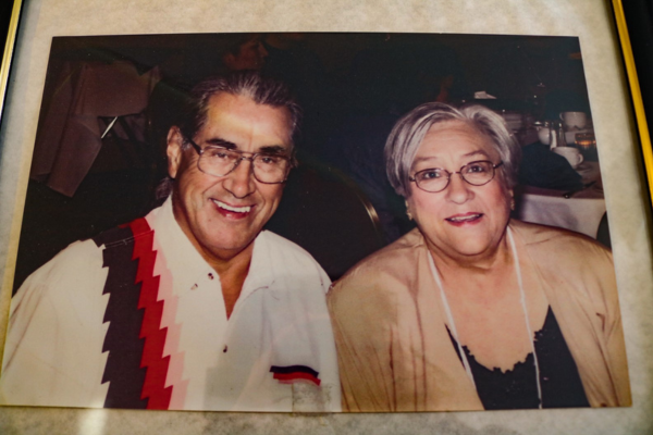 A photograph of a man wearing a white dress shirt with red white and yellow pattern on one side and a woman with short grey hair wearing a tan sweater and pearl necklace