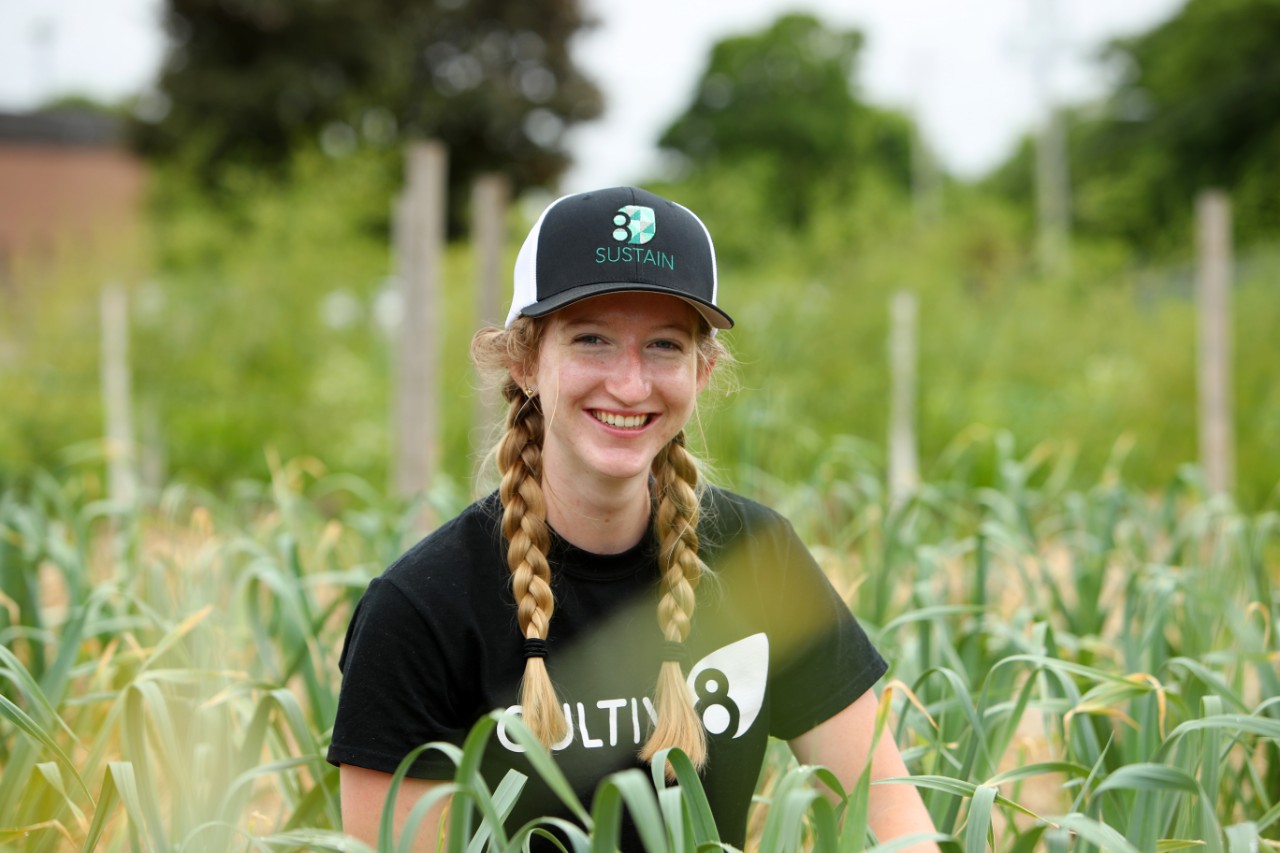 A woman with her hair in two braids and wearing a black ball cap and t-shirt sits in a field of plants