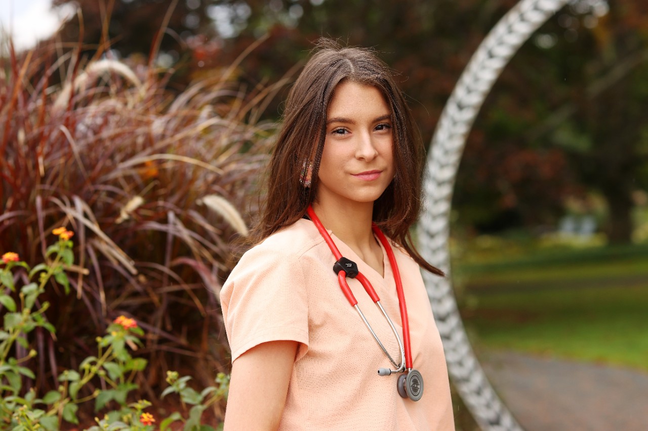 A woman wearing peach-coloured scrubs and a red stethoscope standing in a garden
