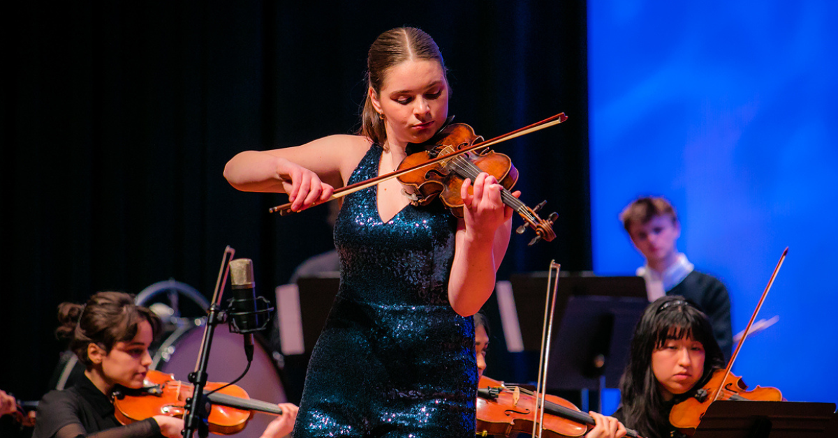 A woman wearing a blue sequined dress plays a violin on stage with other violinists