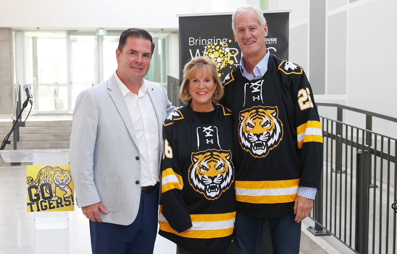 A man dressed business casual stands with a man and a woman wearing black and gold jerseys with a tiger on the front.