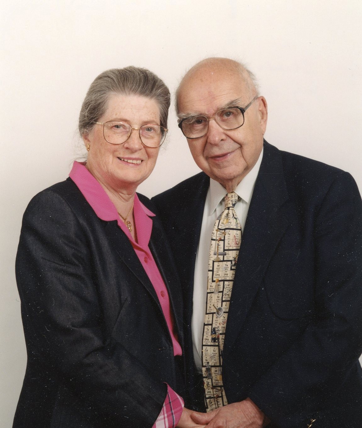 A woman in glasses wearing a pink shirt and dark jacket poses with a man wearing glasses and a suit