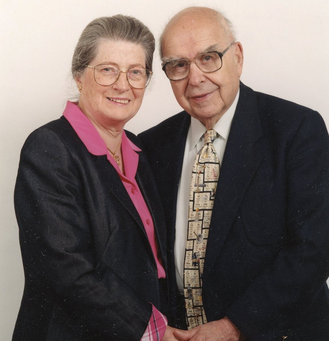 A woman in glasses wearing a pink shirt and dark jacket poses with a man wearing glasses and a suit