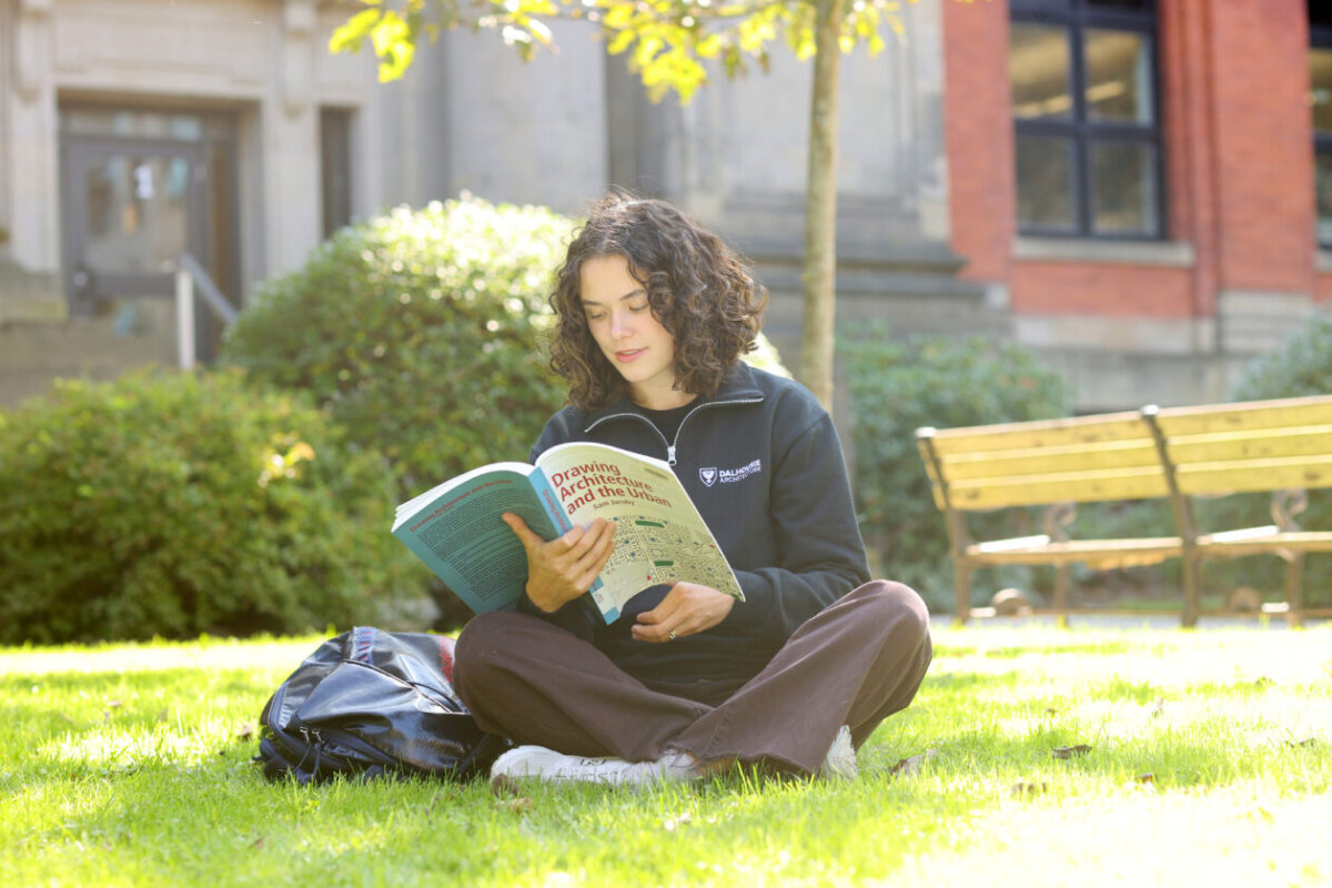 A woman sits cross-legged on the grass reading a book on architecture
