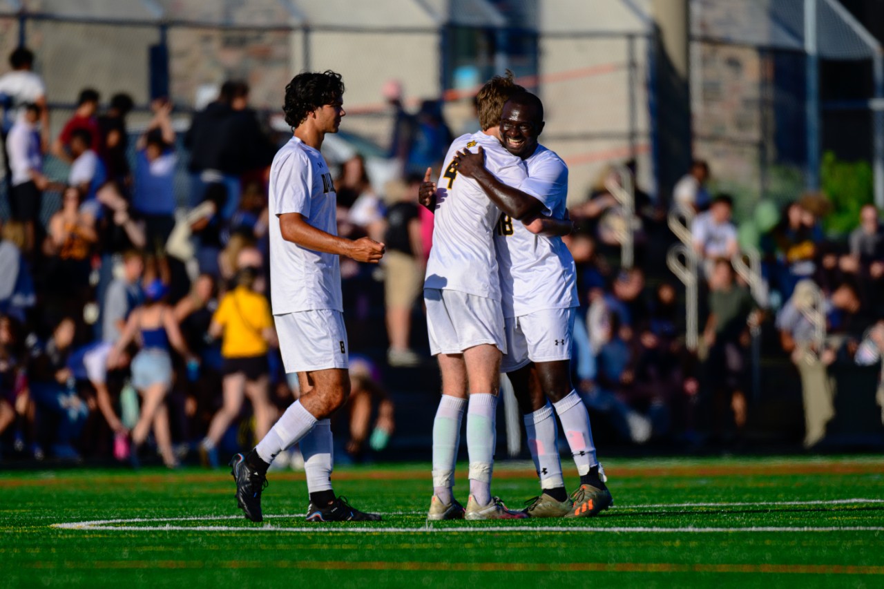 Two men in white soccer uniforms hug while a third man approaches