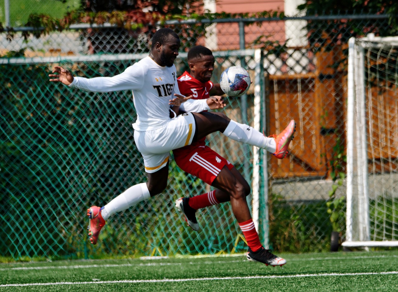 A man wearing a white soccer uniform jumps to intercept the ball from a man in a red soccer uniform