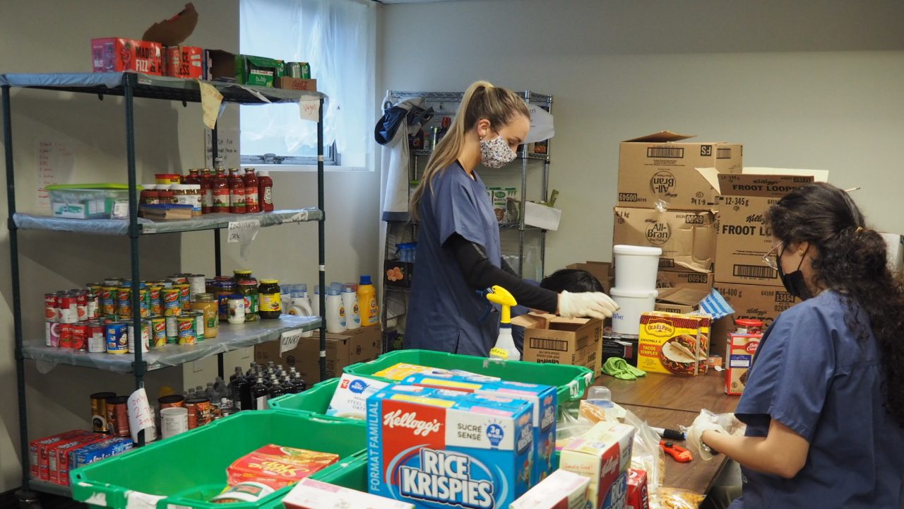 Two people wearing masks and blue tops sort food items at a long table into green bins and onto shelves.