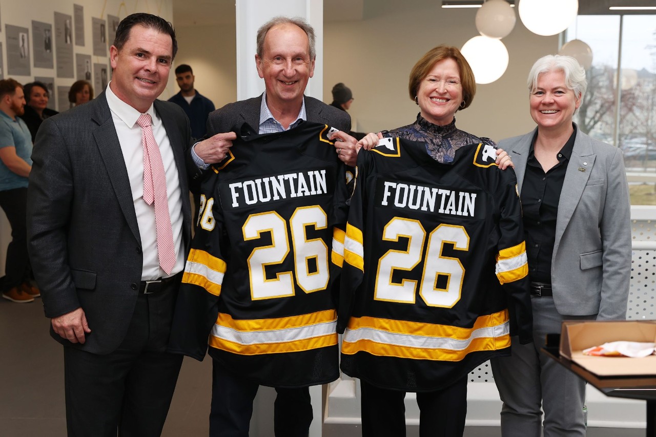 Four people stand together smiling in a lobby at an event with the middle two holding up hockey jerseys.