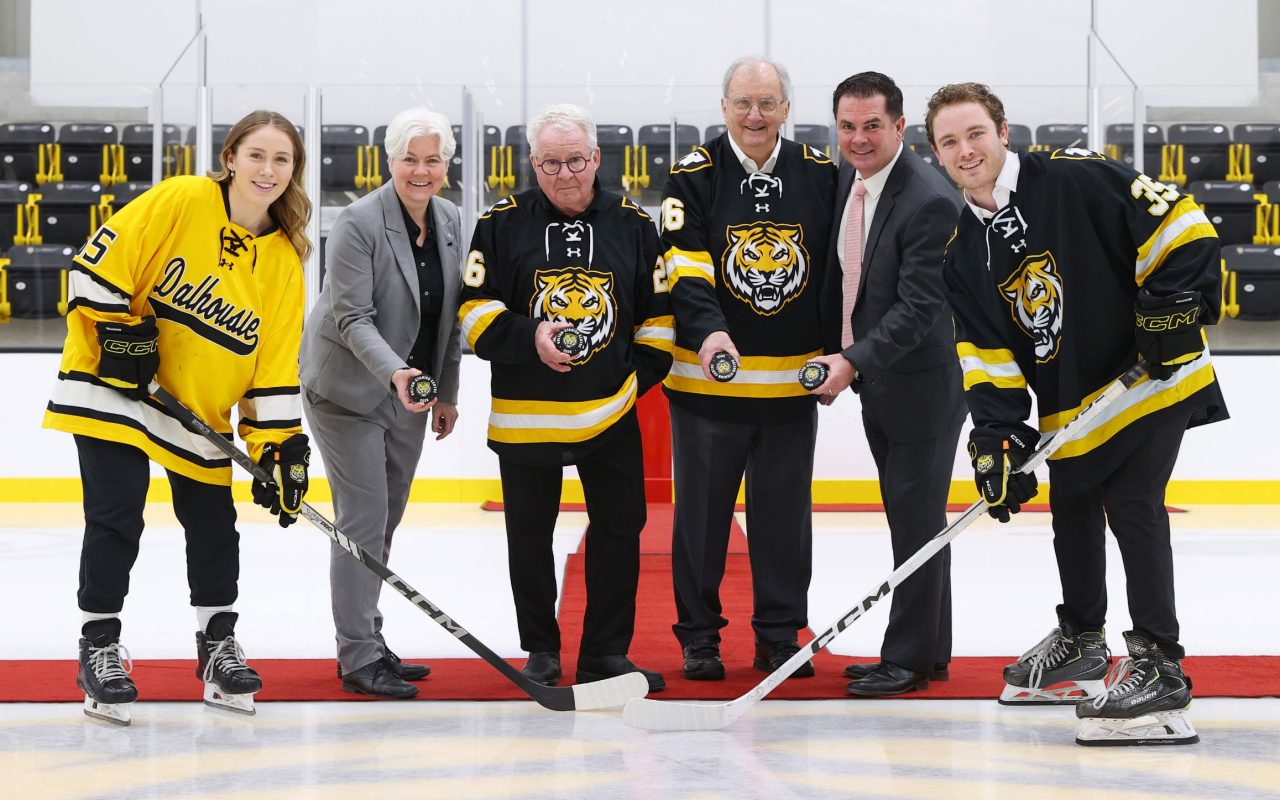 Six people stand on the ice inside a hockey arena: two students in jerseys holding hockey sticks flank four individuals holding pucks, two wearing jerseys and two in suits.