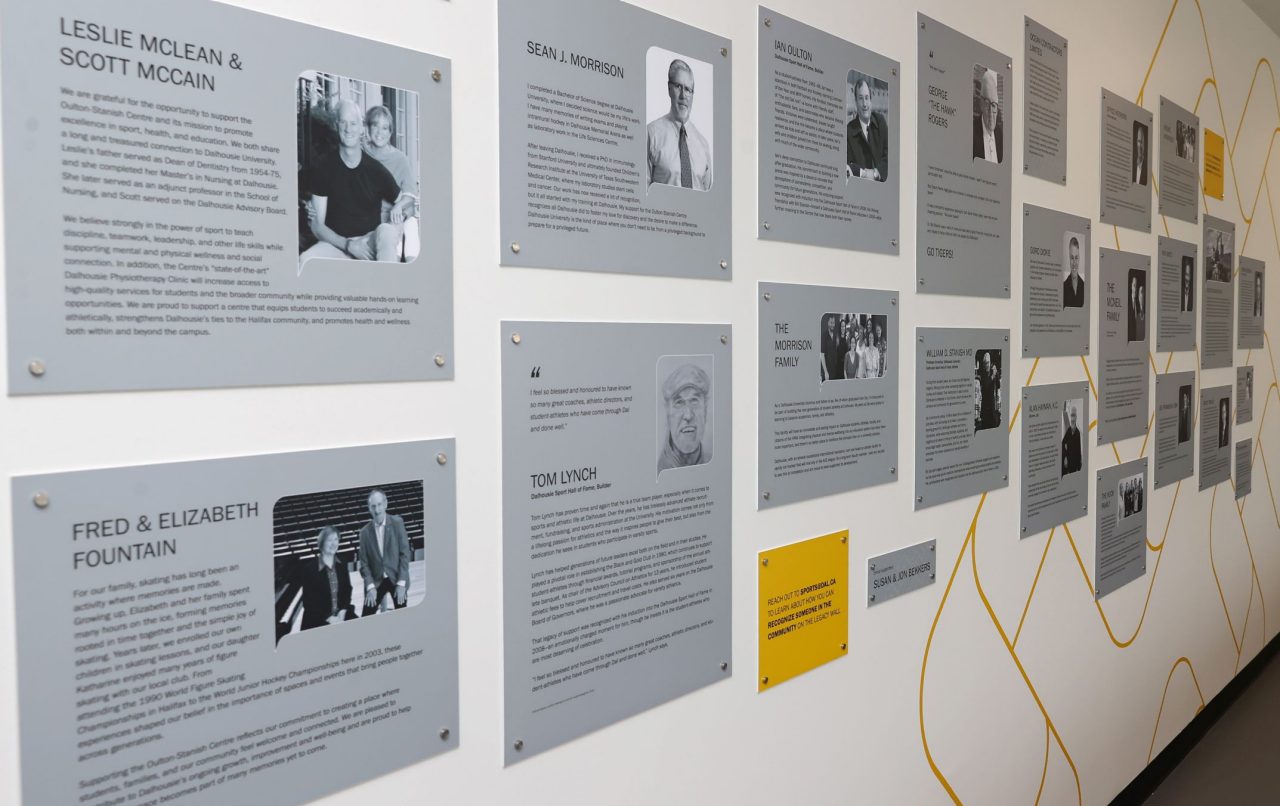 Looking across a white wall installed with many metal plaques with photos of donors and write-ups.