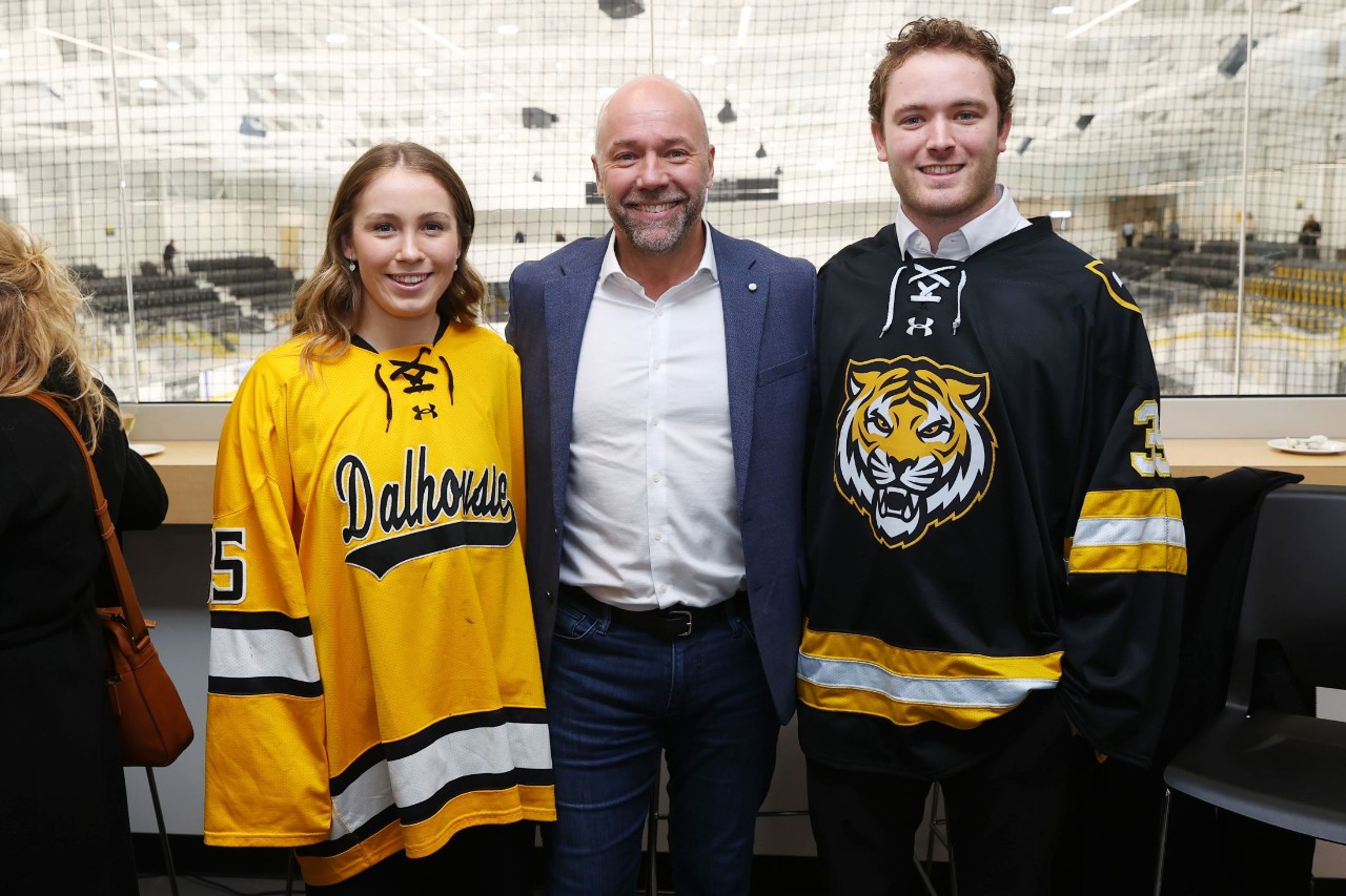A woman and a man in hockey jerseys stand smiling on either side of a man in a suit.