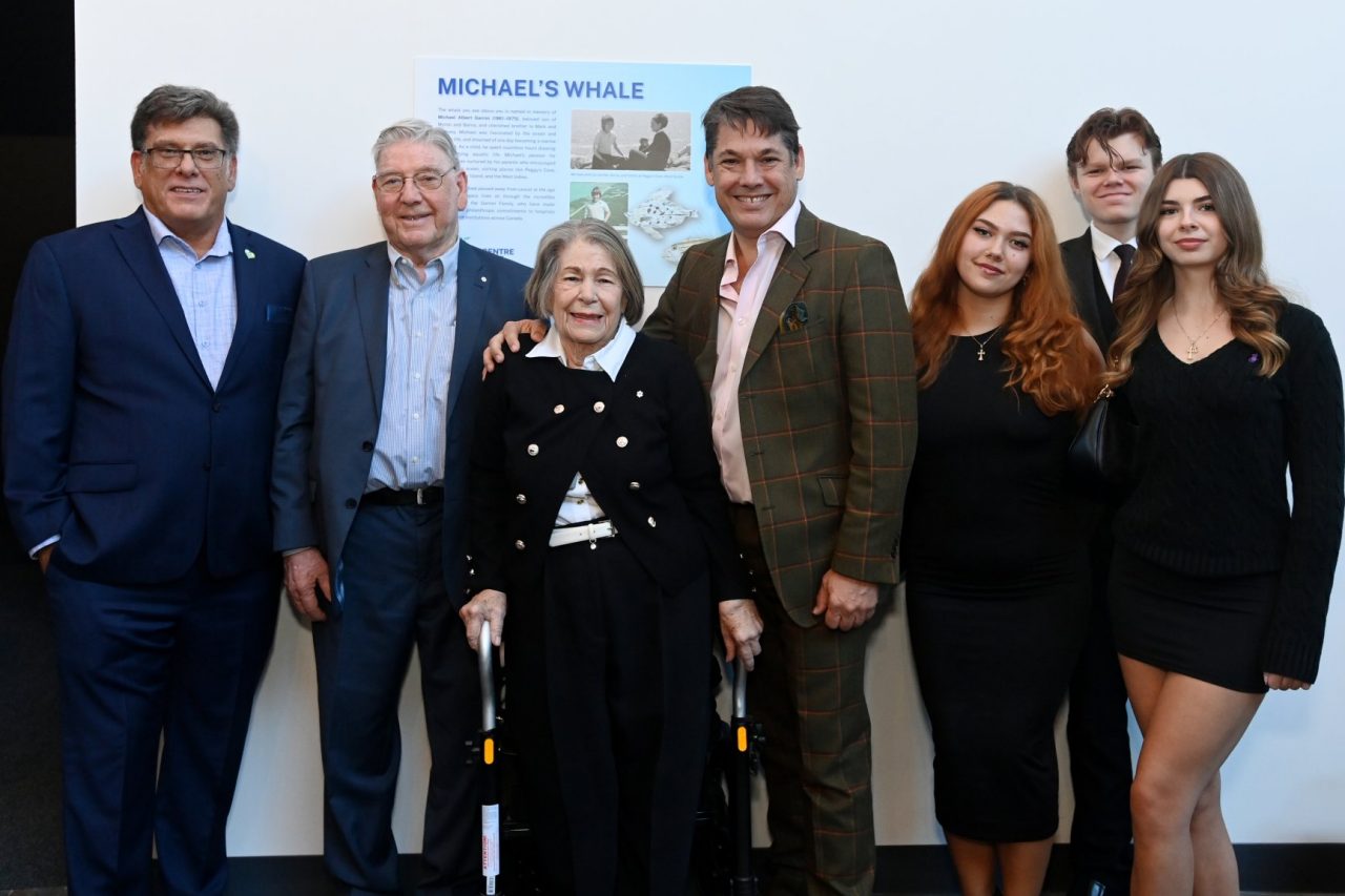 A group of seven people across generations dressed in business casual attire stand together at an event indoors.