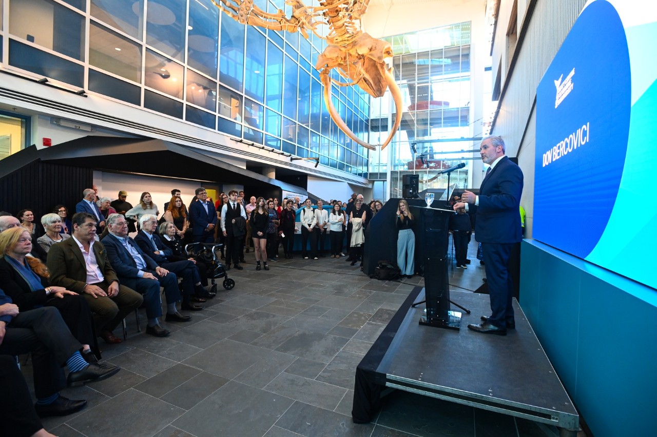 A man speaks at a podium standing on a platform to a group seated and standing in an atrium with the skeleton of a blue whale hanging from the ceiling partially visible. 
