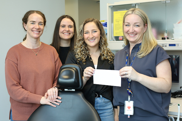 Four women stand around a dental chair holding a cheque