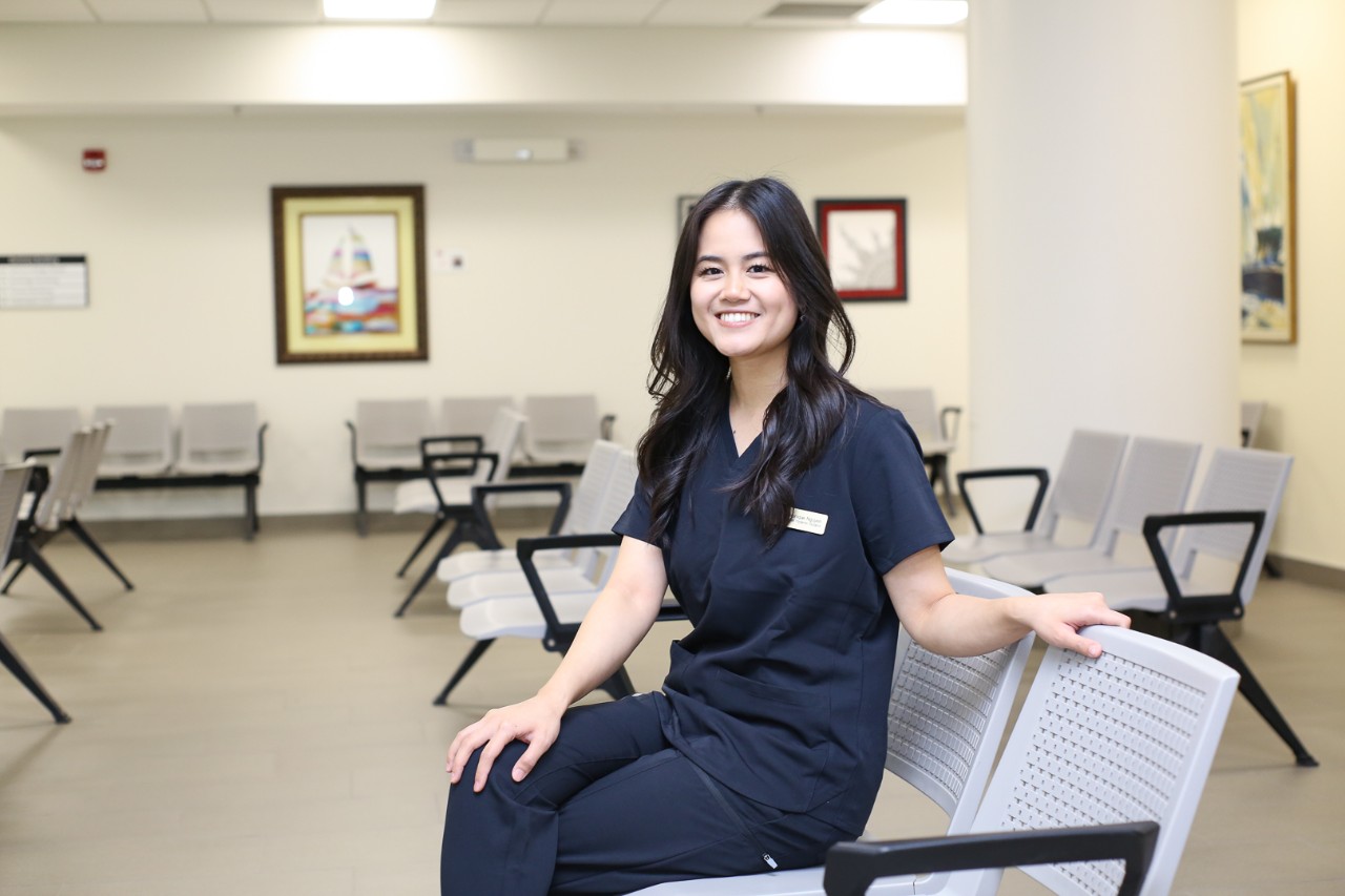 A woman with long black hair and wearing dark blue scrubs sits on a chair in a waiting room