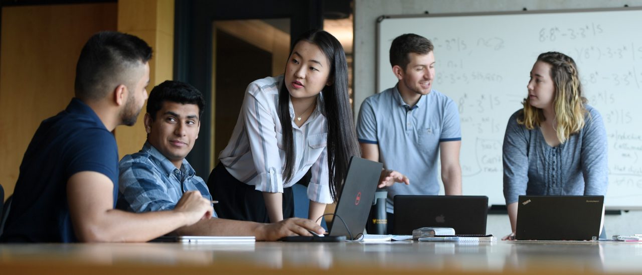A group of young professionals are gathered around a table in conversation, two are seated and three are standing, with laptops open and a whiteboard behind them.