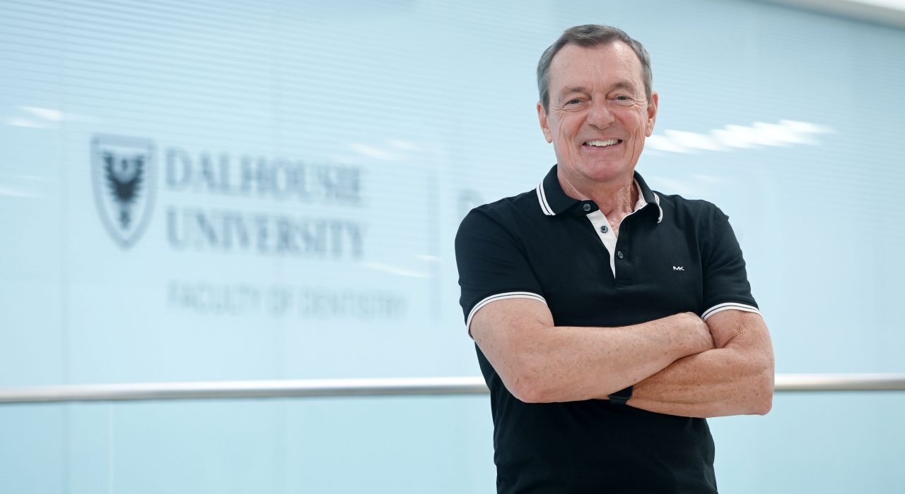 Boran in a black short-sleeved collared shirt standing with his arms crossed smiling in the hallway of the dentistry building.