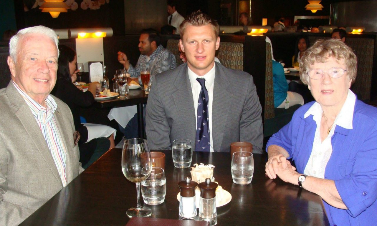 A younger man sits with a man and a woman, all dressed business casually, at a table with glasses on it in a restaurant.