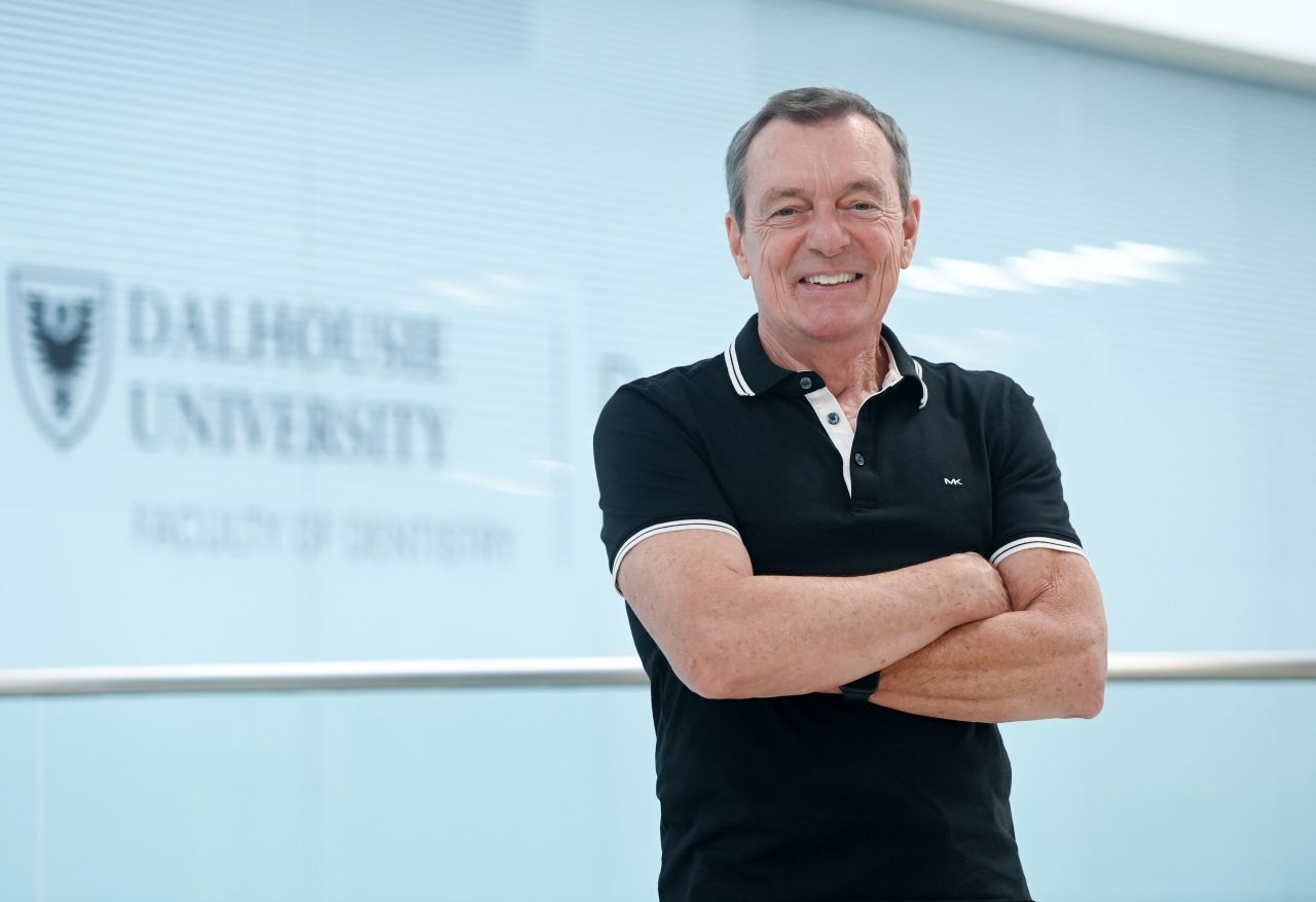 Boran in a black short-sleeved collared shirt standing with his arms crossed smiling in the hallway of the dentistry building.