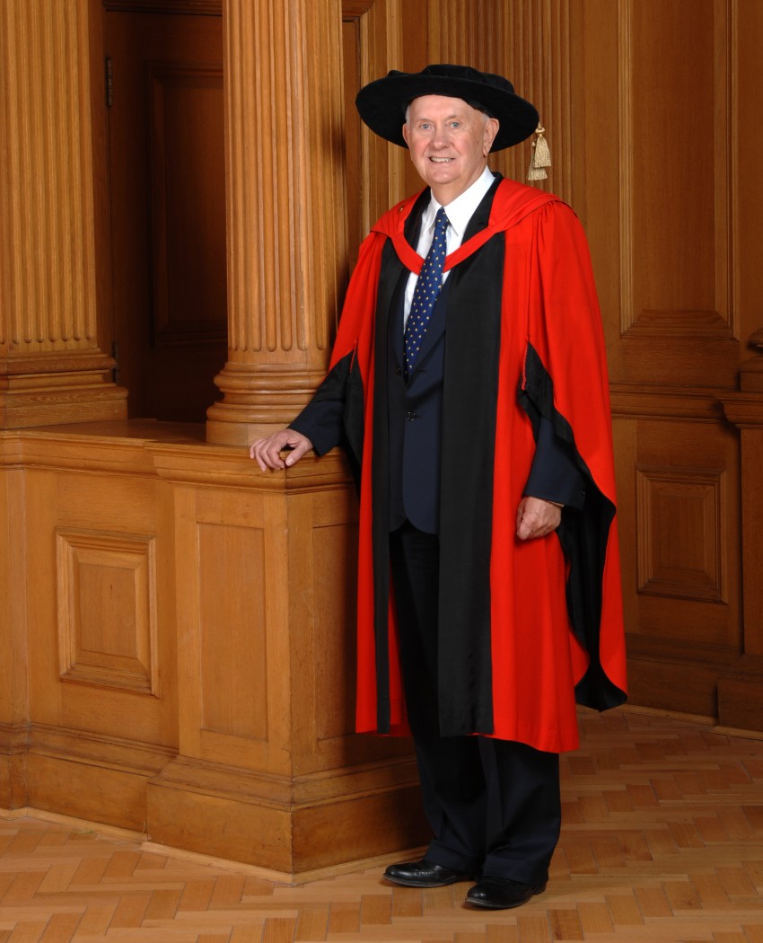 A man wearign a set of red graduation robes poses in a room with wooden finishes