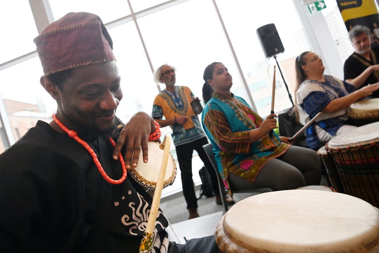 A group of people playing various drums indoors, dressed in colorful attire, with a large window in the background.