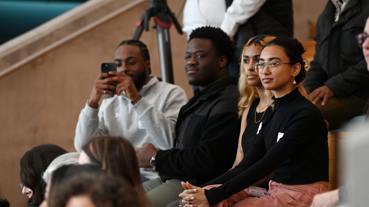 A group of diverse attendees sit on bleachers indoors at an event while one records on his phone.