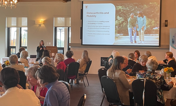 People sitting and dining at round tables at an indoor venue with a large screen displaying slides.