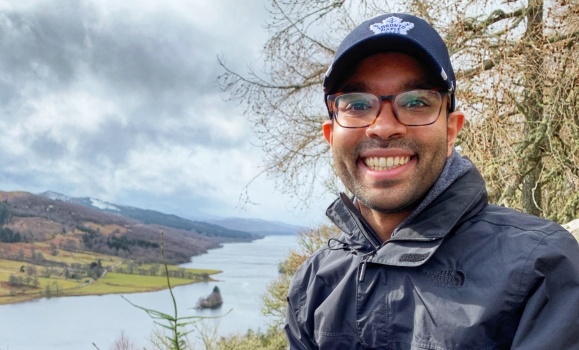 A man wearing a jacket and baseball cap with a view of a river and fields in the back ground