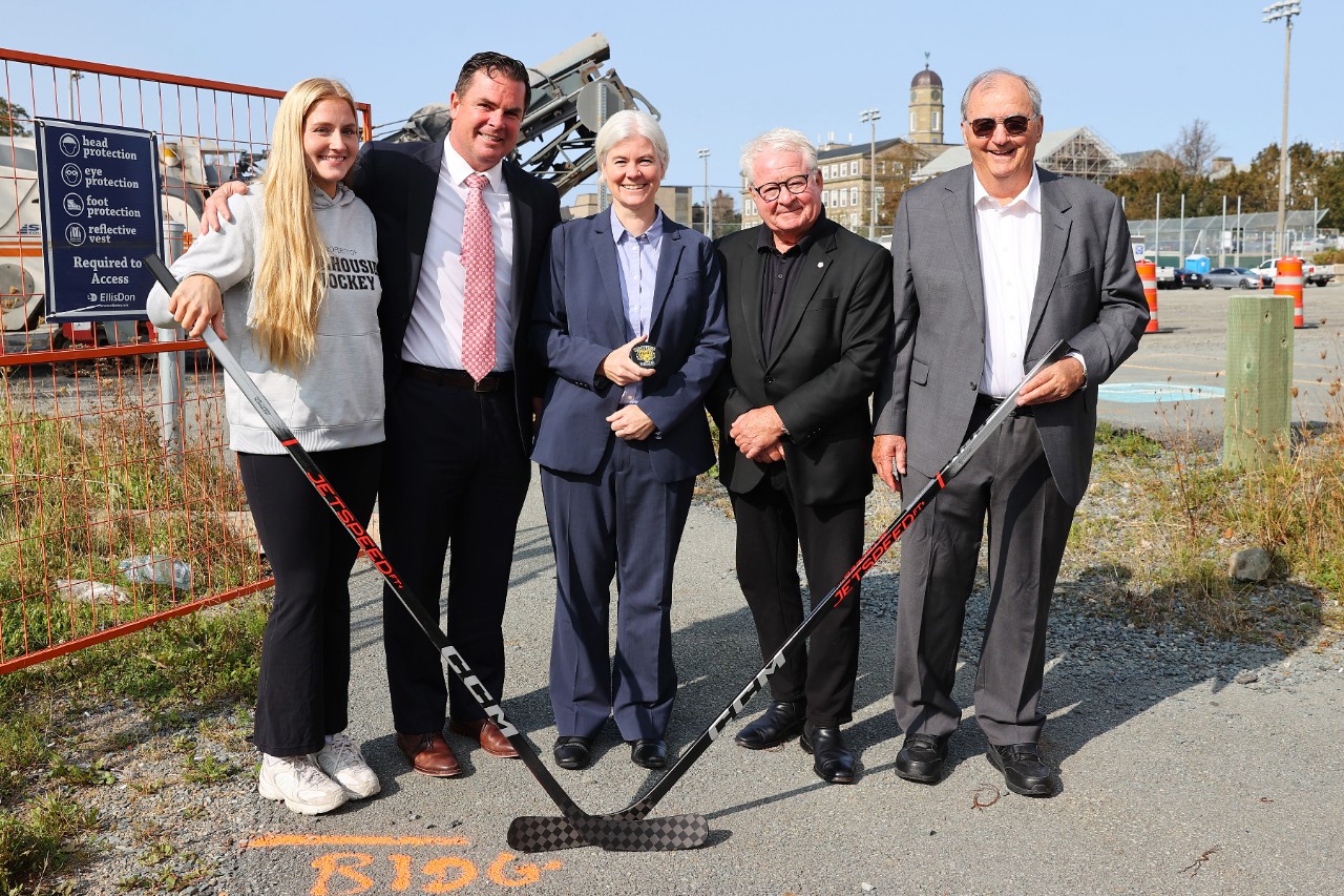 A group of men and women holding hockey sticks pose in front of a construction site