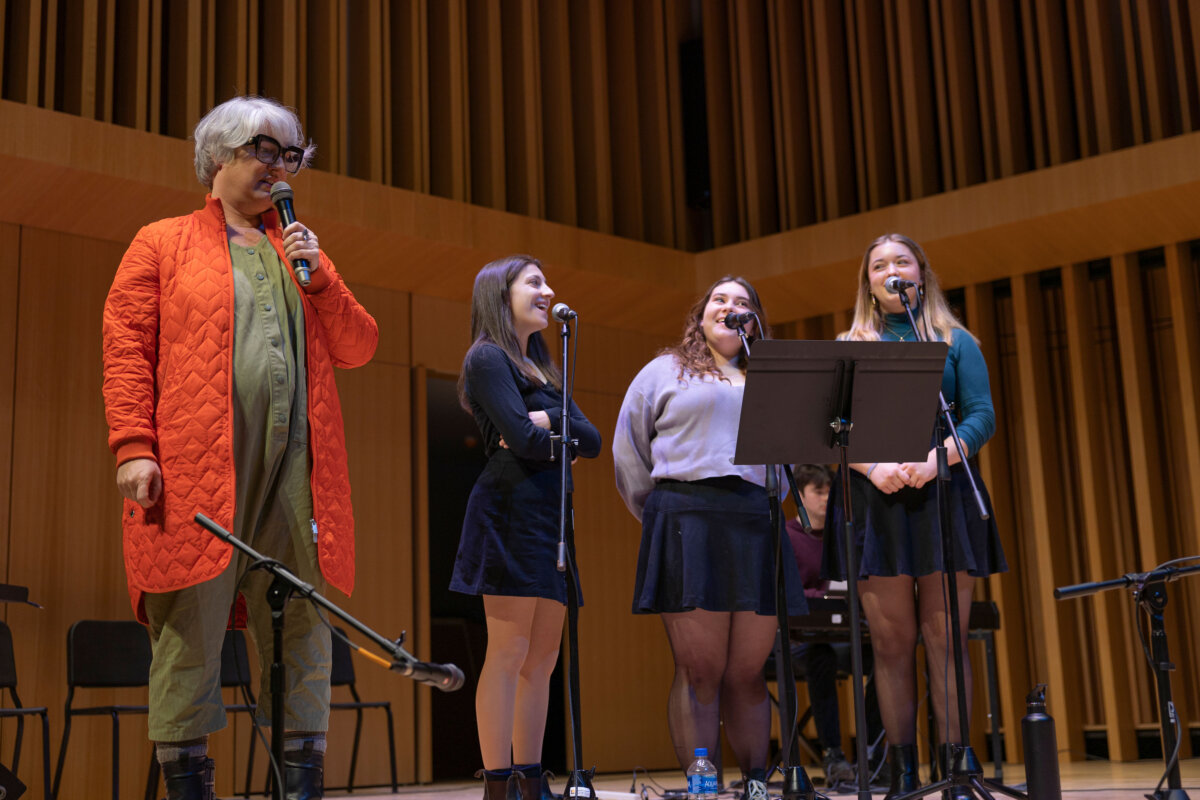 A woman in an orange jacket stands on stage with three singers