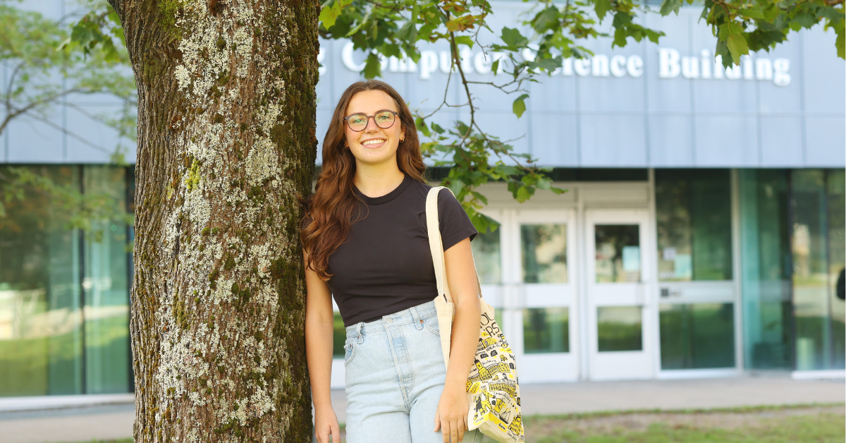 A woman wearing glasses and a black t-shirt and bag leans against a tree in front of the Goldberg Computer Science building