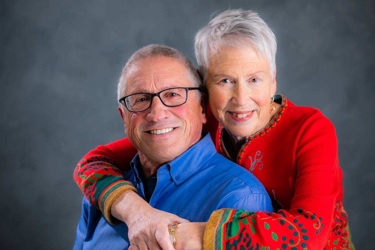 A woman with short grey hair wraps her arms around a man in a blue shirt.