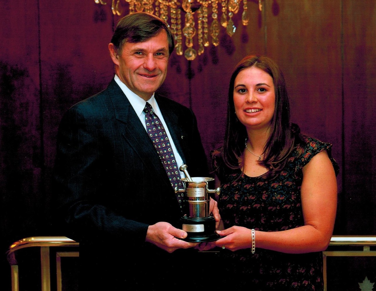 A man wearing a suit and tie holds a small trophy with a woman wearing a floral print blouse