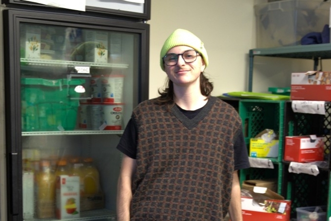 Holman leaning on a table in front of a tall fridge and crates of supplies in the food bank.