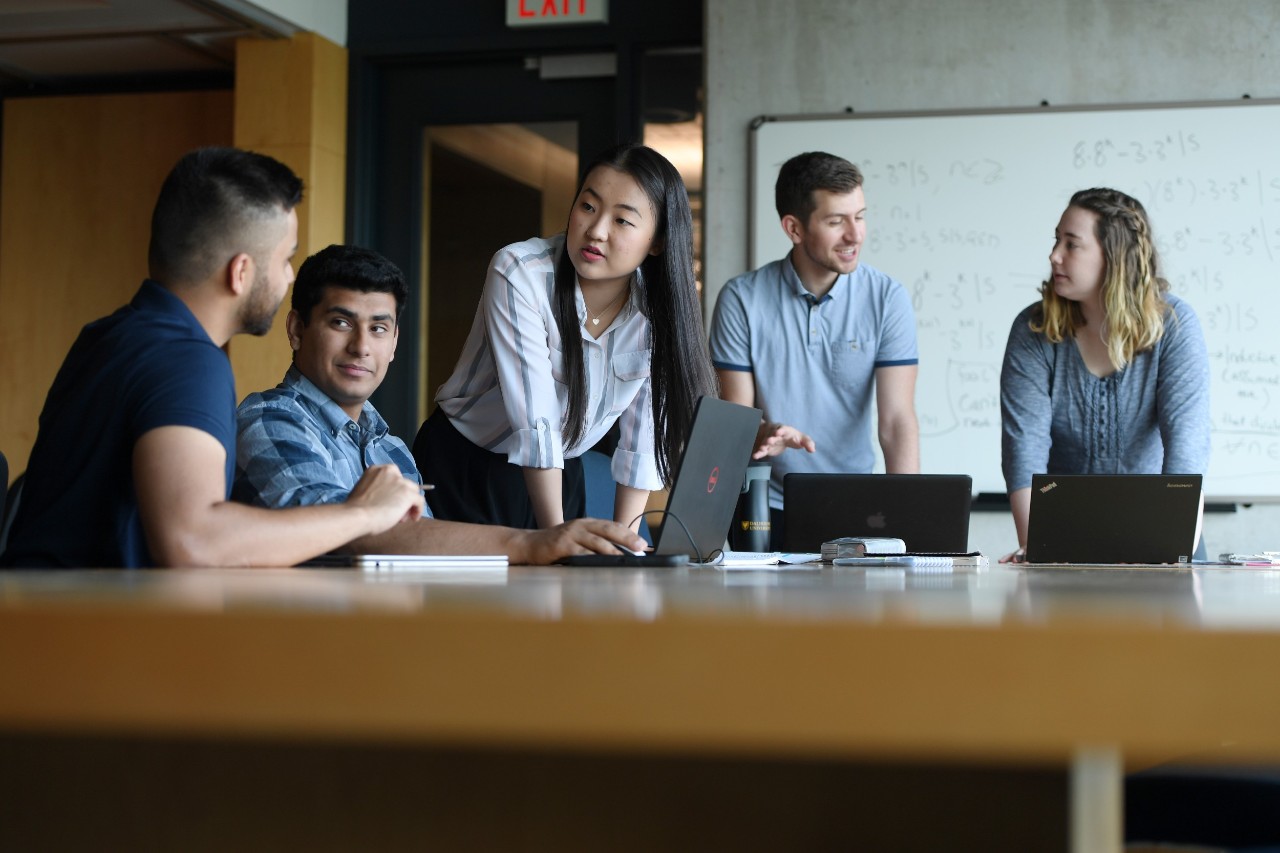 A group of young professionals are gathered around a table in conversation, two are seated and three are standing, with laptops open and a whiteboard behind them.