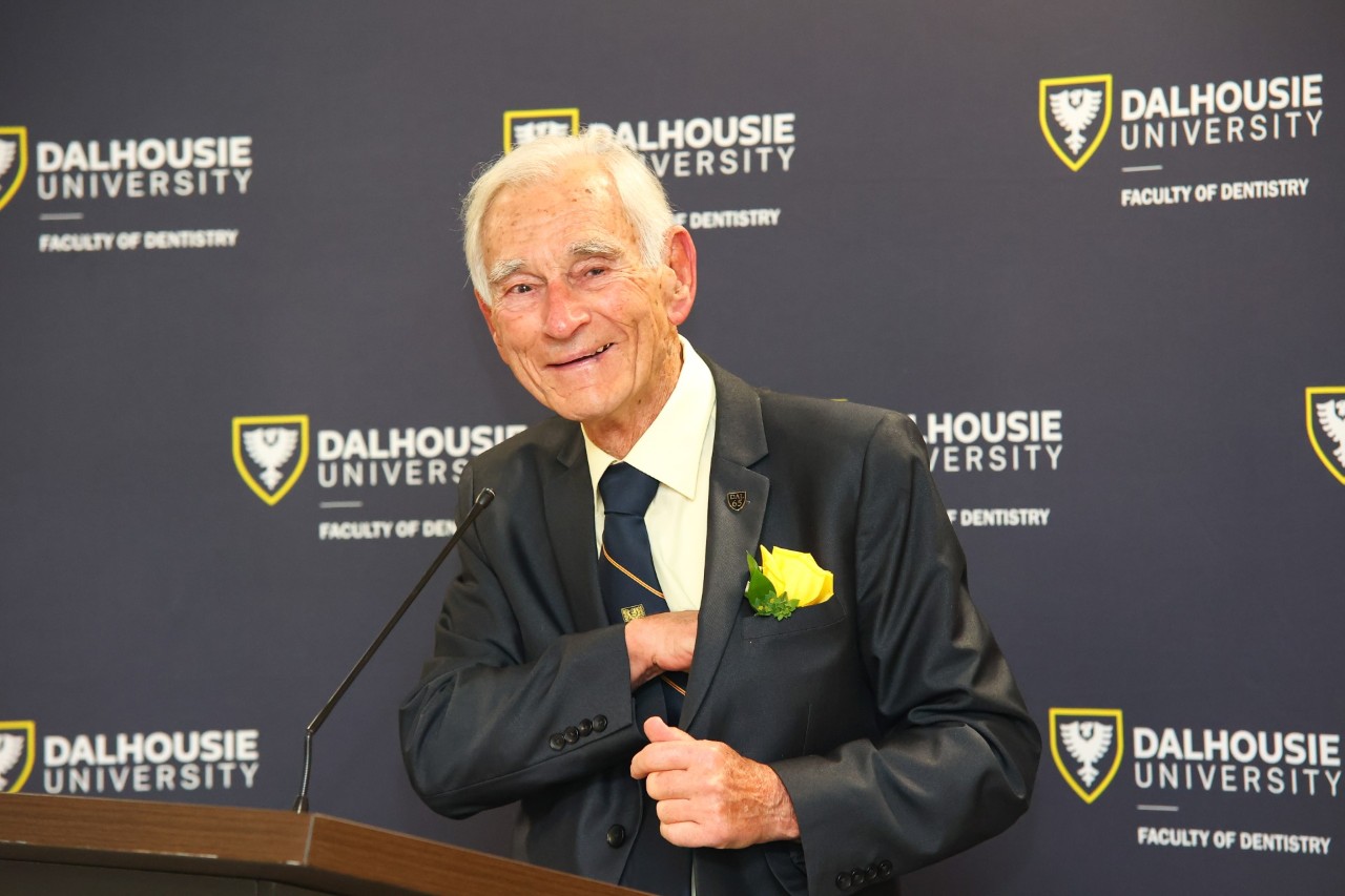 Frank Lovely stands at a podium dressed formally preparing to speak in front of a backdrop of Faculty of Dentistry logos.