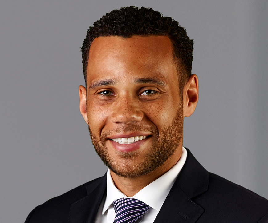 A headshot of a man wearing a black suit and blue and purple striped tie