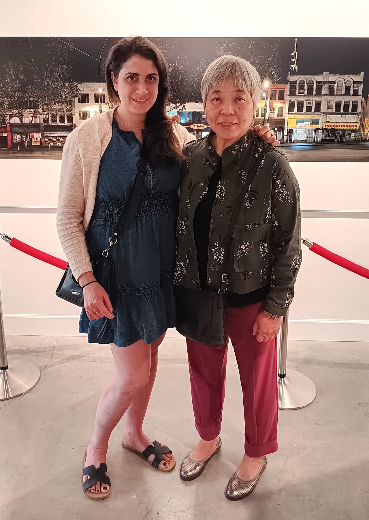 Two woman dressed casually stand together indoors in front of a photograph of a streetscape.