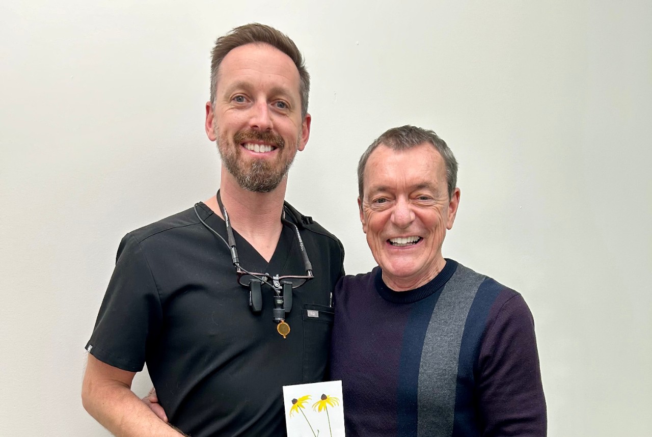 A man wearing black scrubs stands alongside a man in a purple sweater who is holding a card with two yellow yellow flowers.