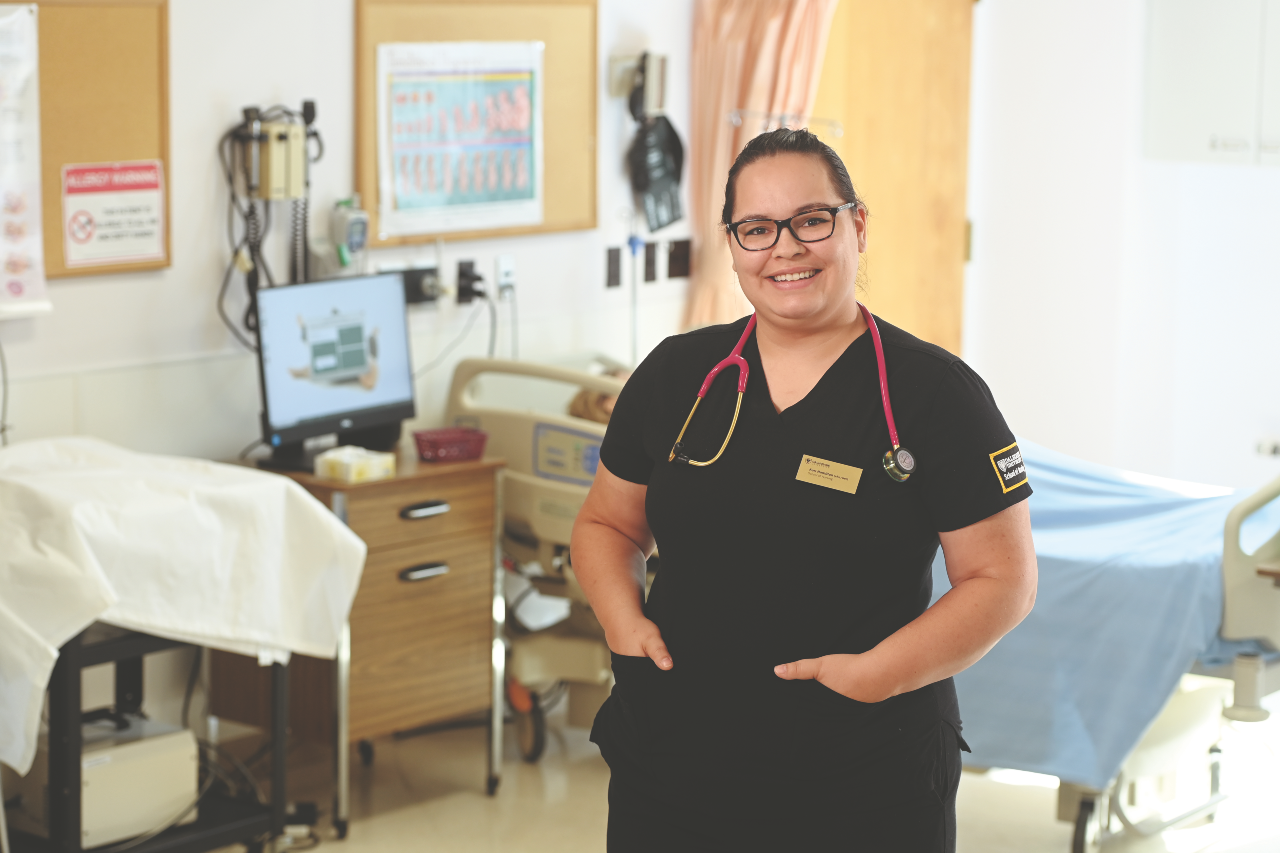 A woman wearing glasses and black scrubs with a pink stethoscope standing in a hospital room