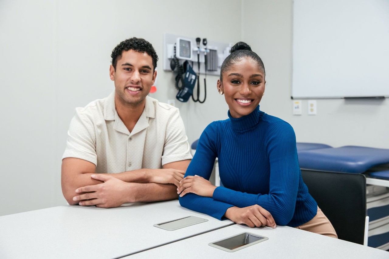 A male-presenting and a female-presenting student sit together both with arms folded on a table, smiling in a medical exam room. 
